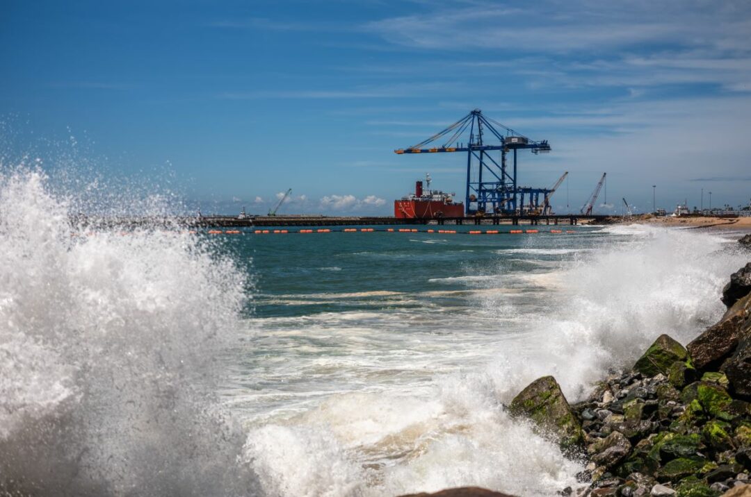 A PORT WITH CRANES IS SEEN IN THE DISTANCE, ACROSS A STRETCH OF SEA, WITH WAVES CRASHING ON ROCKS IN THE FOREGROUND