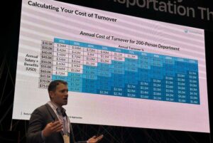 A MAN IN A JACKET HOLDING A MICROPHONE SPEAKS IN FRONT OF A LARGE SCREEN SHOWING A CHART
