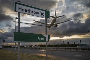 A green sign alongside a road that reads "Heathrow," with a white passenger airplane flying overhead