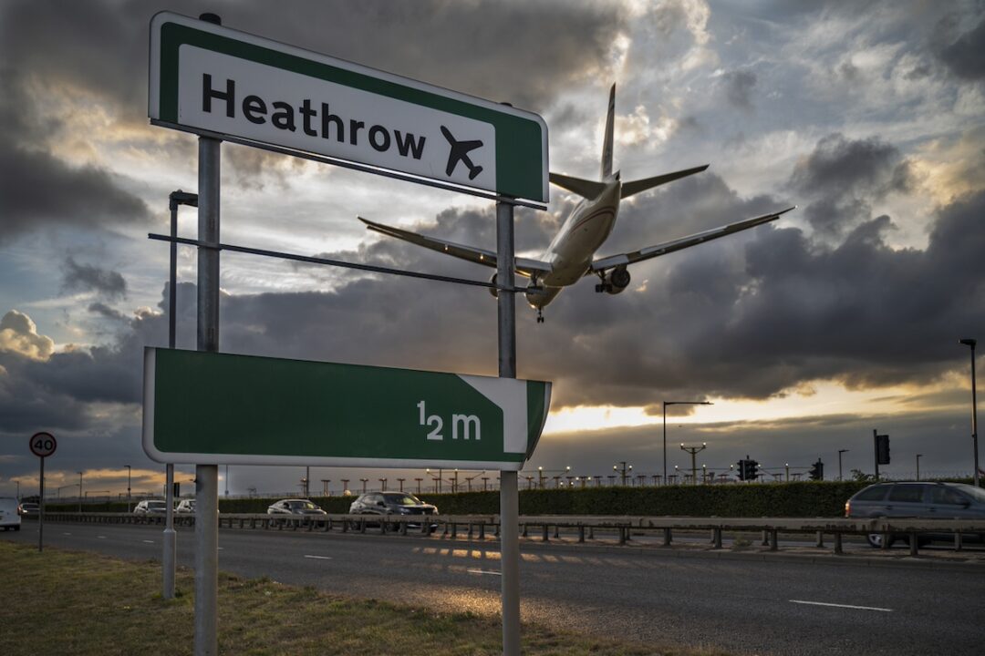 A green sign alongside a road that reads "Heathrow," with a white passenger airplane flying overhead