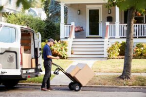 A DELIVERY WORKER WITH A HAND TRUCK LOADED WITH BOXES OUTSIDE A VAN PARKED IN FRONT OF A HOME CONSULTS A DEVICE