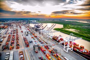 A seaport at sunset with rows of multi-colored shipping containers stacked in rows, with white shipping cranes lining the shoreline