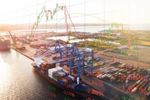 A black container ship stacked with containers docked under two blue cranes, with a green and red line graph superimposed over the skyline