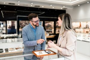 A MAN AND A WOMAN SMILE AND TALK OVER ITEMS OF JEWELRY ON TOP OF A DISPLAY CASE IN A STORE