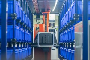 BLUE PLASTIC CRATES SIT IN MULTI-ROW SHELVING IN A WAREHOUSE, A YELLOW ROBOTIC MACHINE VISIBLE AT THE END OF THE AISLE